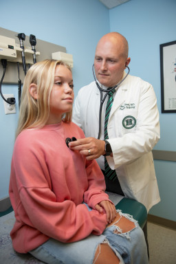 A white man in a doctor's coat listens to a young white girl's heart with a stethoscope.