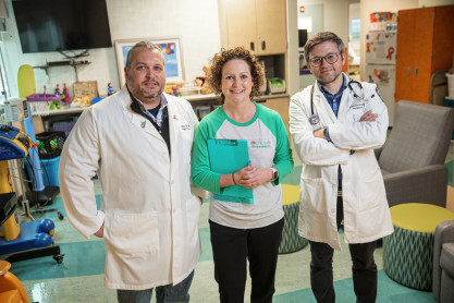Two male doctors in white coats pose for a photo with a female nurse practitioner wearing a green and white T-shirt holding a clip board. The background is a hospital room where children receive infusions. 