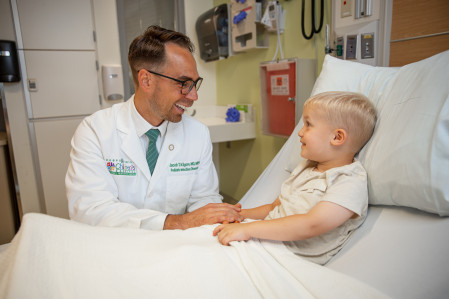 A male doctor in a white coat holds the hand of a young boy lying in a hospital bed. They are both smiling.